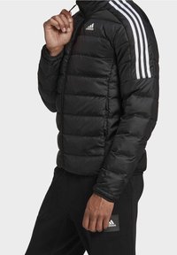 Homme portant une veste matelassée noire Adidas et un pantalon noir, avec trois rayures blanches sur les manches de la veste et le logo Adidas sur la poitrine et le pantalon.