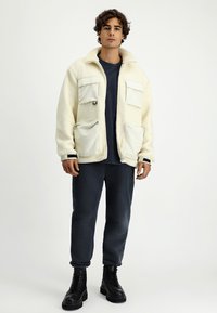 Beżowa polarowa kurtka z czterema przednimi kieszeniami, wysokim kołnierzem i zamkiem. Noszona na granatowej koszuli, w połączeniu z granatowymi dresami i czarnymi butami.