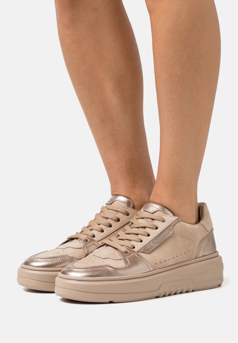 Baskets beiges avec des accents métalliques portées par une personne, montrées de la partie inférieure du mollet vers le bas sur un fond neutre.