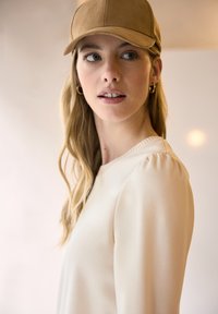 Jasnobeżowa bluzka o gładkiej fakturze z marszczeniem na ramionach, w połączeniu z jasnobrązową czapką z daszkiem, na tle o miękkim odcieniu.