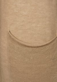 Beżowa dzianina ma teksturowaną powierzchnię z widoczną kieszenią. Projekt jest prosty, z zaokrąglonym brzegiem kieszeni i miękkim wyglądem.