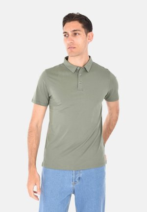 Uomo con polo a maniche corte verde oliva e jeans azzurri, in piedi con una mano dietro la schiena, che guarda di lato su sfondo bianco.