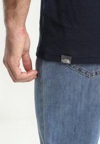 Námořnické bavlněné tričko s prošitým lemem, na kterém je vidět logo od The North Face. Nosené s jeansy světle modré barvy, ruka spočívající na tričku.