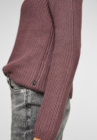 Sweter dzianinowy w kolorze przykurzonego różu z teksturowanym wzorem. Posiada metkę po boku i długie rękawy, zestawiony z szarymi dżinsami.