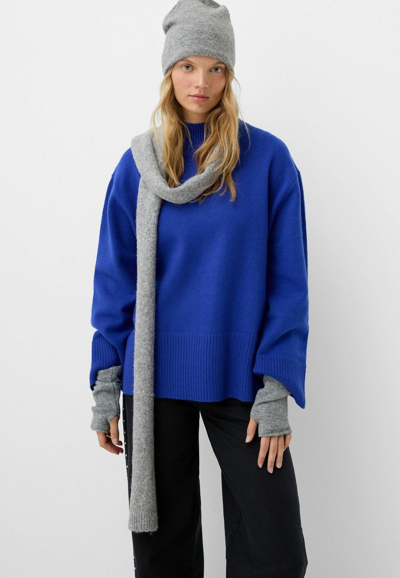 Pull en tricot bleu avec ourlet côtelé, associé à une écharpe grise et un bonnet assorti. Le pull présente des manches larges et des trous pour les pouces.
