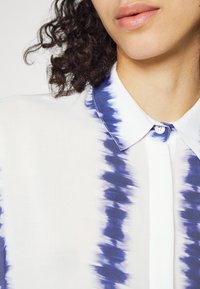 Camicia colletto in tessuto bianco trasparente con motivo tie-dye blu navy. Presenta una chiusura con bottoni e un orlo arrotondato. Texture liscia con una leggera lucentezza.