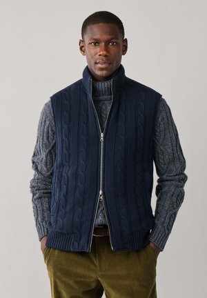 Hombre con chaleco de punto de canalé azul marino con cremallera sobre un suéter de punto de canalé gris y pantalones de pana verde oliva.