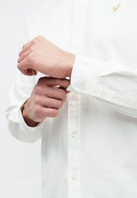 Biela košeľa na gombíky s hladkou textúrou, s dlhými rukávmi a malým vyšívaným logom na hrudi. Gombíky sú viditeľné.