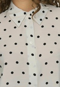 Korte mouw, lichtgekleurde stof shirt met een knoopsluiting aan de voorkant, voorzien van grote zwarte stippen en een spitse kraag. Glad texture.