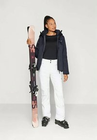 Skikleding met een houten ski met zwarte en roze accenten, gecombineerd met een waterdichte marineblauwe jas, zwart shirt en witte skibroek.