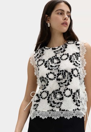 Kvinde iført hvid ærmeløs blondertop med sorte blomsterprint og bølgede kanter, der kigger til siden mod en ensfarvet baggrund.