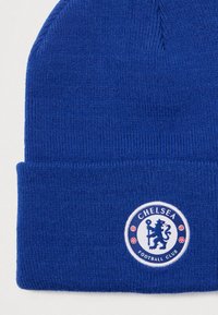 Zils adīts cepure ar salocītu malu, kurā ir apaļa Čelsi futbola kluba uzlīme baltā krāsā ar lauvu emblemām un sarkanām akcentiem.