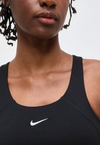 Κοντινό πλάνο στο κάτω μέρος του προσώπου και του λαιμού ενός ατόμου που φοράει μαύρο αθλητικό σουτιέν Nike με λευκό λογότυπο Swoosh στο στήθος, με απλό φόντο.