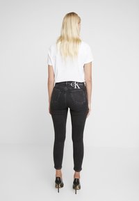Svarta skinny jeans med hög midja, med fickor och en CK-logotyp på midjebandet, kombinerat med en vit t-shirt med kort ärm och svarta klackar.