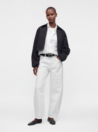 Personne portant une veste courte noire, une chemise blanche boutonnée, un pantalon blanc avec une ceinture noire et des ballerines noires, debout devant un fond uni.