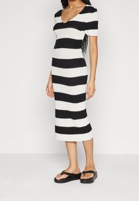 Gestreiftes Strickkleid in Schwarz und Weiß, mit V-Ausschnitt und kurzen Ärmeln, eng anliegender Silhouette und knielangem Design. Getragen mit schwarzen Flip-Flops.