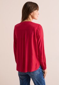 Blusa rossa a maniche lunghe con vestibilità morbida, realizzata in tessuto liscio. Presenta un alto yoke posteriore e fessure laterali, abbinata a jeans in denim.
