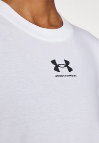 Bijela pamučna majica s okruglim izrezom. Na prsima se nalazi mali crni Under Armour logo, a tkanina je glatka i mekana.