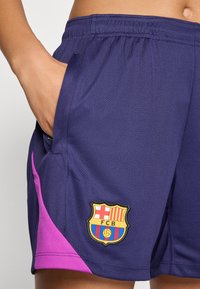 Shorts de sport violets avec des accents roses, arborant un logo FC Barcelone brodé. Conçus en tissu léger et texturé.
