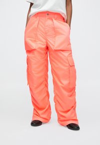 Személy neon narancssárga, széles szárú, több zsebes cargo nadrágot visel, fekete slip-on cipővel, deréktól lefelé látható.