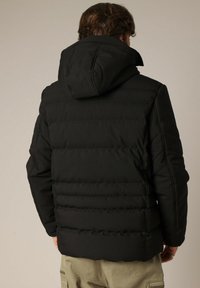 Man in een zwarte gewatteerde jas met capuchon, gezien van achteren, gecombineerd met khaki cargobroek tegen een effen achtergrond.