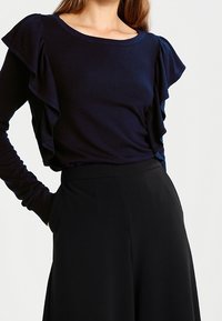 Top lungo blu navy con maniche a strati con dettagli a volant, abbinato a pantaloni neri a vita alta. Tessuto morbido e liscio.