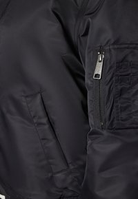 Zwarte bomberjack met een gladde, glanzende textuur, met een ritszak op de mouw en zijzakken voor opbergruimte.