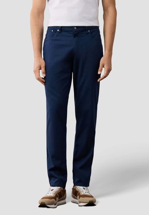 Man die marineblauwe rechte broek, wit T-shirt en bruine en witte sneakers draagt, staand tegen een effen achtergrond.