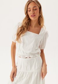 Blusa di cotone bianca con maniche corte a palloncino e scollatura quadrata, con motivi a occhiello floreali e orlo arricciato. Indossata con una gonna abbinata.