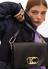 En kvinna med långt hår håller en svart läderhandväska med en guldlogo, klädd i en marinblå puffjacka och en lila topp, ser med självförtroende in i kameran.