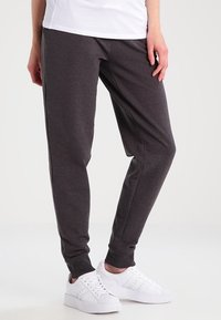 Persoon die donkergrijze joggingsbroek, witte sneakers en een wit shirt draagt, staand tegen een effen lichte achtergrond.