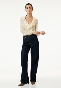 Creme bluse med flæser ved udskæringen og lange ærmer, parret med mørkeblå, brede denimbukser og lysebrune spidse hæle.
