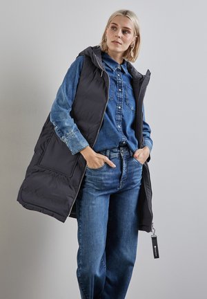Gilet rembourré noir avec capuche sur une chemise en denim bleu, associé à un jean bleu. Le gilet a une poche frontale et des détails de fermeture éclair argentés.