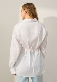 White button-up shirt with a tied waist, long sleeves, and a slightly curved hem. Smooth fabric with a relaxed fit and classic collar.