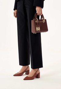 Hnědá kožená kabelka se zlatou kovovou výbavou a logem, doplněná o hnědé boty na blokovém podpatku se špičatou špičkou a černé připravené kalhoty.