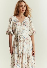 Blomstret kjole med V-hals og puffede ærmer, featuring en indsnævret talje og et lagdelt skørt. Farverne inkluderer hvide, lyserøde og blå blomster på en lys baggrund.