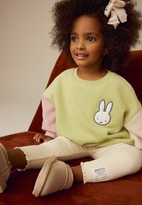 Dvodijelna sweatshirt u zelenoj i ružičastoj boji s grafikom zeca, rebraste kreme hlače i svijetle papuče. Dijete s kovrčavom kosom sjedi na smeđoj stolici.