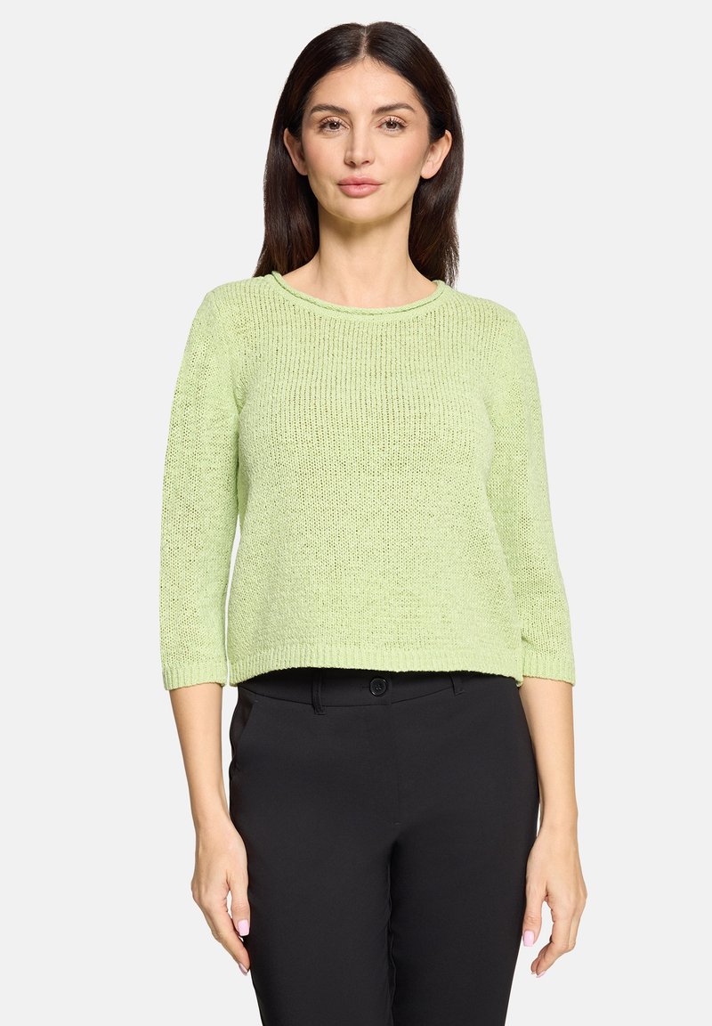 Femme portant un pull en tricot vert clair à manches trois-quarts et un pantalon noir, debout devant un fond clair uni.
