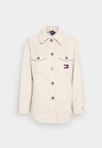 Kremowa koszula-zszywka z mieszanki wełny z kołnierzykiem, zapinana na guziki, z dwiema kieszeniami na piersi oraz naszywką z logo. Posiada metalowe guziki oraz zaokrąglony dół.