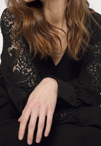 Žena s vlnitými hnědými vlasy, oblečená v černých krajkových šatech a zlatém řetízku, s levou rukou uvolněně nataženou vpřed.
