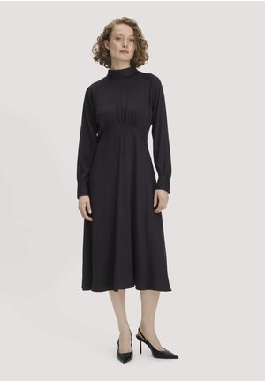 Vrouw met krullend haar, gekleed in een zwarte midi-jurk met lange mouwen en zwarte puntige hoge hakken, staand op een effen lichte achtergrond.