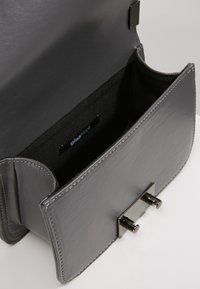 Šedá kožená kabelka s hladkou texturou, vybavená zapínáním na sponu a jednoduchou vnitřní podšívkou. Uvnitř je viditelná malá značka značky.