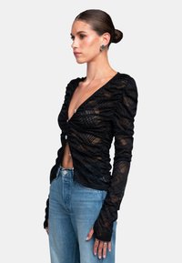 Schwarzes Langarmoberteil mit gerafften Details, tiefem V-Ausschnitt und einem durchsichtigen, strukturierten Stoff. Trägt man über blauen Jeans.