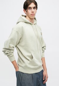 Sudadera verde claro, mezcla de algodón, con bolsillo canguro, capucha con cordón, puños y dobladillo acanalados. Combinada con pantalones sueltos gris oscuro.