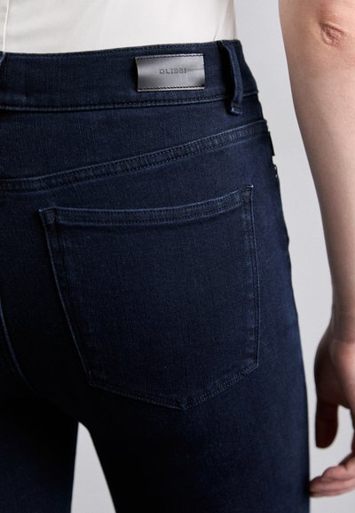 Un jean en denim bleu foncé présente une taille droite avec un patch en cuir, des poches et des détails de couture discrets à l'arrière.