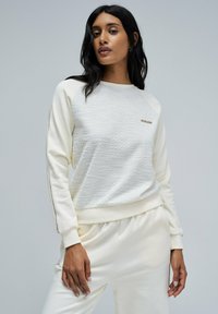 La sudadera crema presenta un frente texturizado, mangas raglán y acentos laterales. Suave tejido y corte relajado mejoran la comodidad.