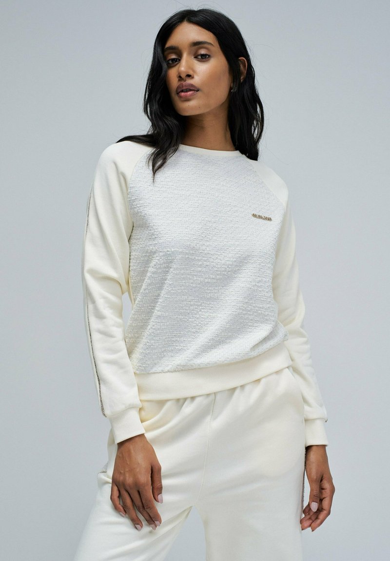La sudadera crema presenta un frente texturizado, mangas raglán y acentos laterales. Suave tejido y corte relajado mejoran la comodidad.
