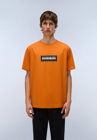 Orange bomulds t-shirt med en sort rektangulær grafik med "NAPAPIJRI geographic." Enkel rund hals og korte ærmer.