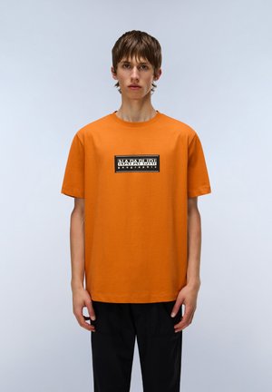 Orange bomulds t-shirt med en sort rektangulær grafik med "NAPAPIJRI geographic." Enkel rund hals og korte ærmer.