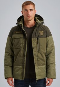 Grön vadderad jacka med huva, försedd med flera fickor, dragkedjor och ett emblem. Tygets textur verkar slät och hållbar.